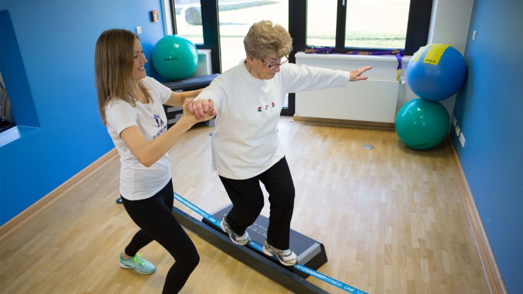 Senior woman training balance on slackline platform with instructor support
